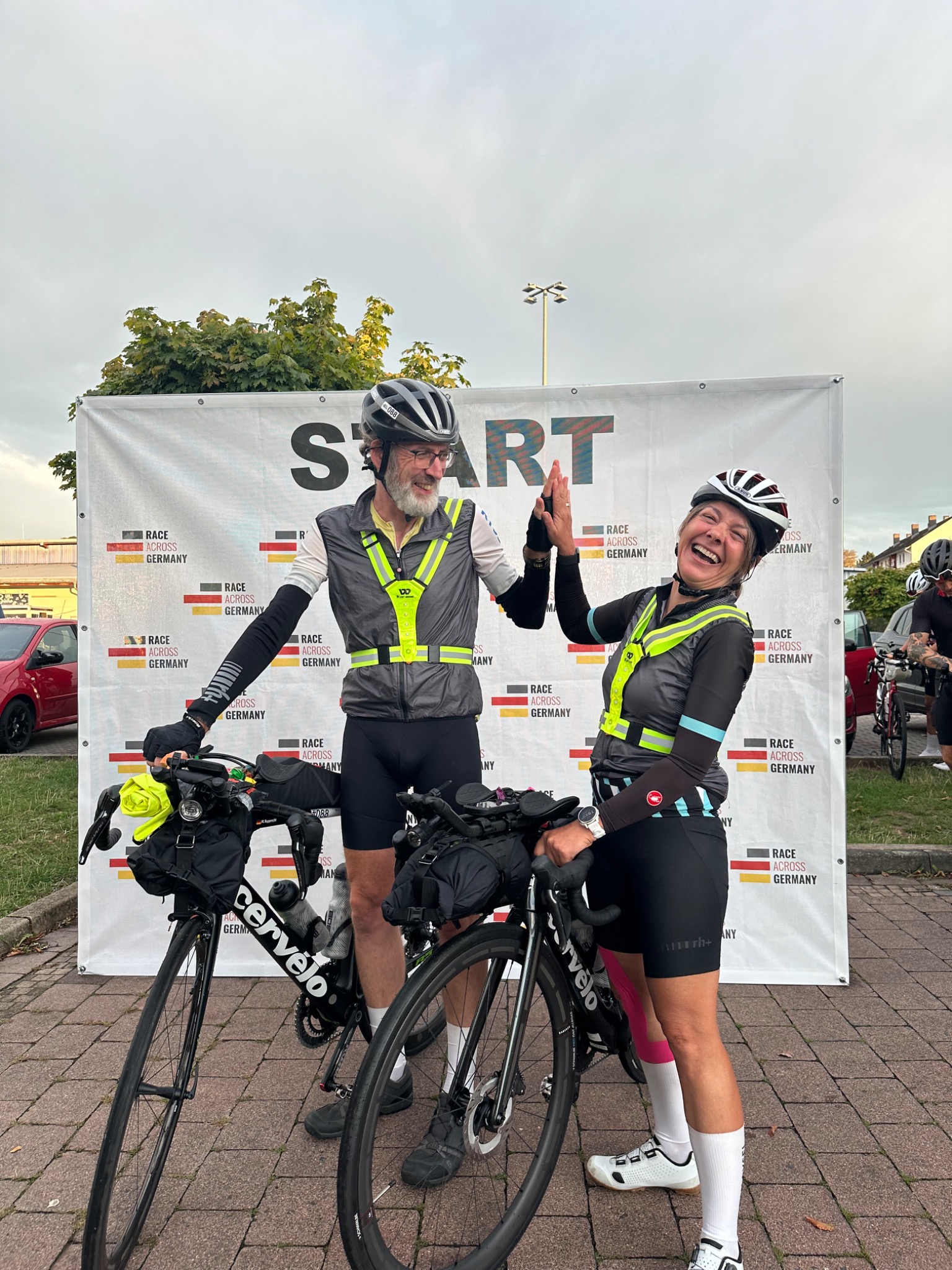 Diana und Marco mit Rennrädern und reflektierenden Westen beim gemeinsamen Abklatschen vor der Race-Across-Germany-Startwand